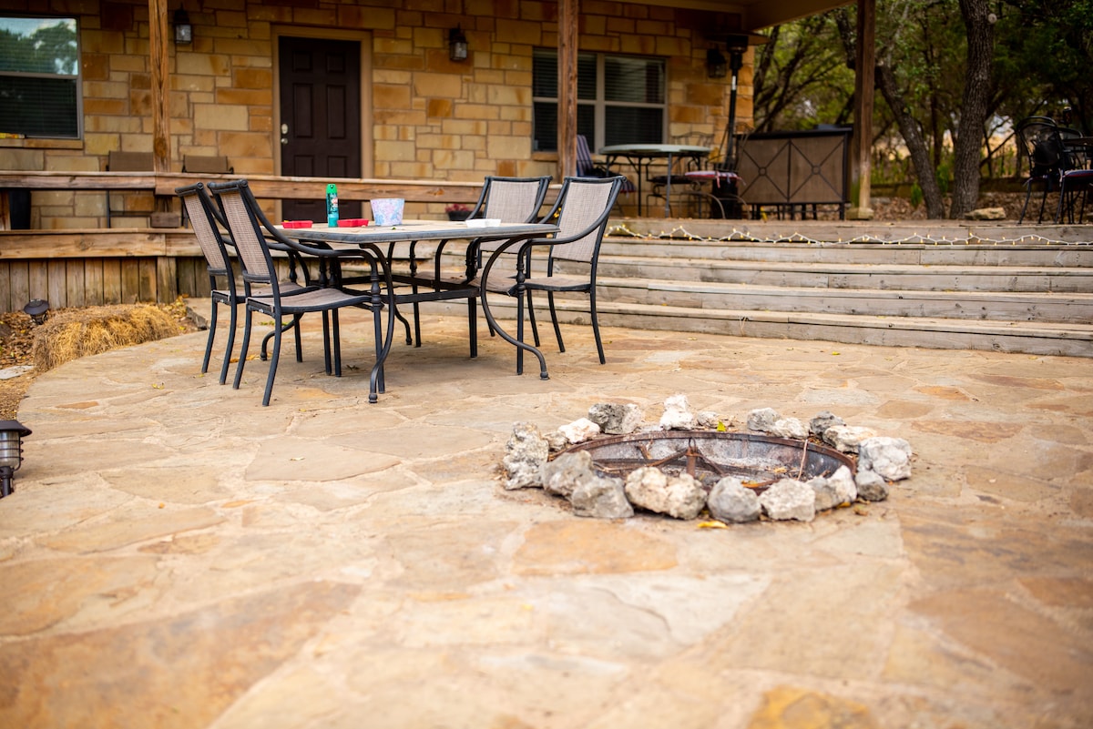 A stone-paved patio features a circular fire pit made of rocks, surrounded by a metal table and six chairs. The area offers access to a wooden deck and showcases the surrounding natural landscape.