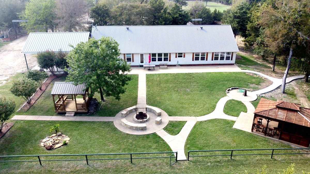 Aerial view of Little Creek Ranch showcases the expansive green lawn surrounding the house. A stone fire pit is positioned near a paved pathway, leading to a gazebo on the left and another covered seating area. The landscape is complemented by trees and open space.