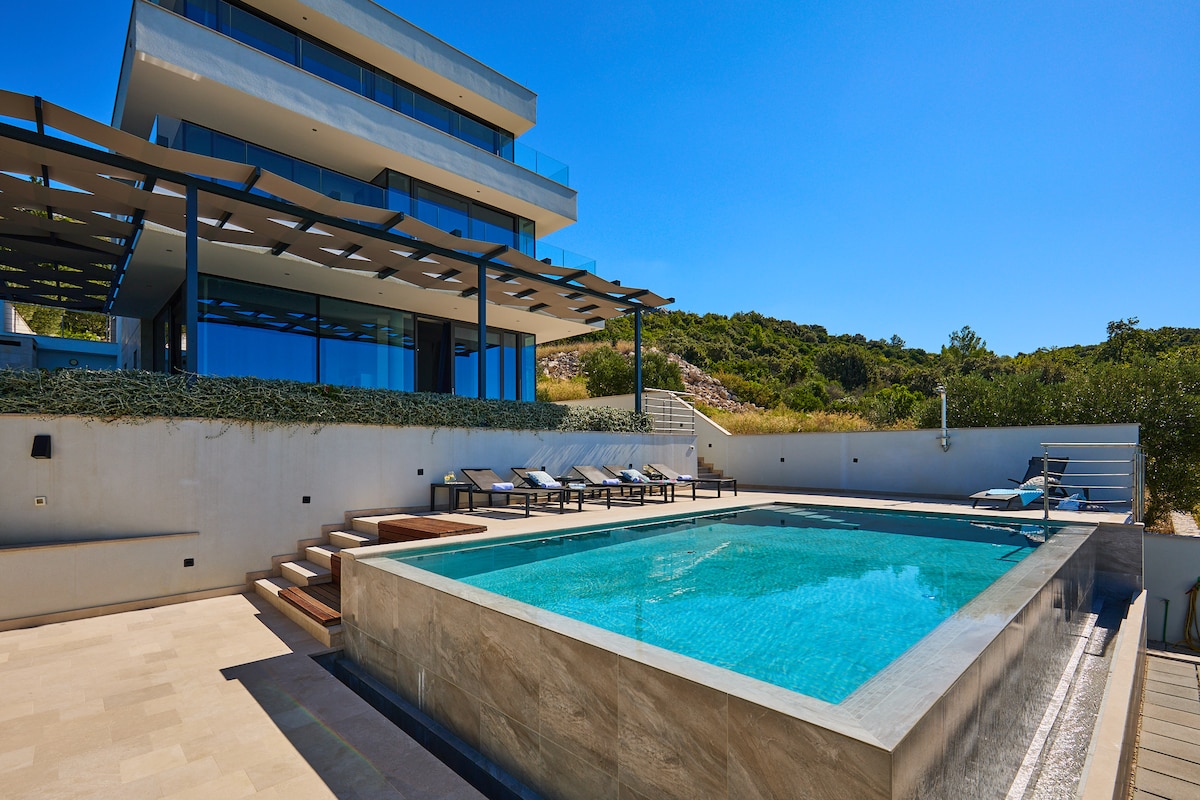 A modern villa is depicted with a private swimming pool set within a spacious terrace area. Sun loungers are arranged around the pool, and expansive glass panels reflect the bright blue sky. Lush greenery borders the property, contributing to a serene outdoor space.