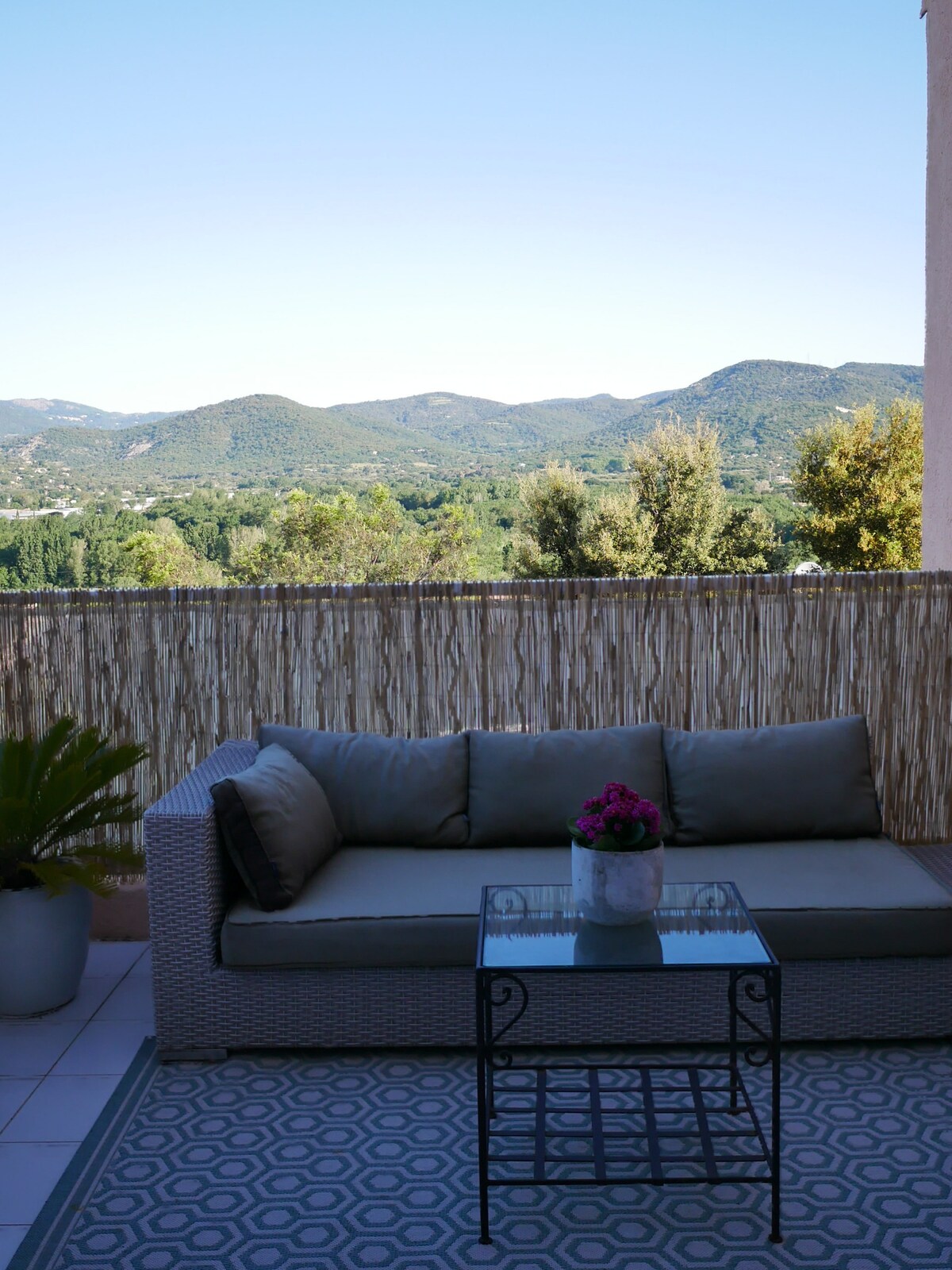 A comfortable outdoor seating area is displayed, featuring a soft sofa and a glass table. The surrounding view overlooks rolling hills and greenery under a clear blue sky, creating a serene backdrop for relaxation.
