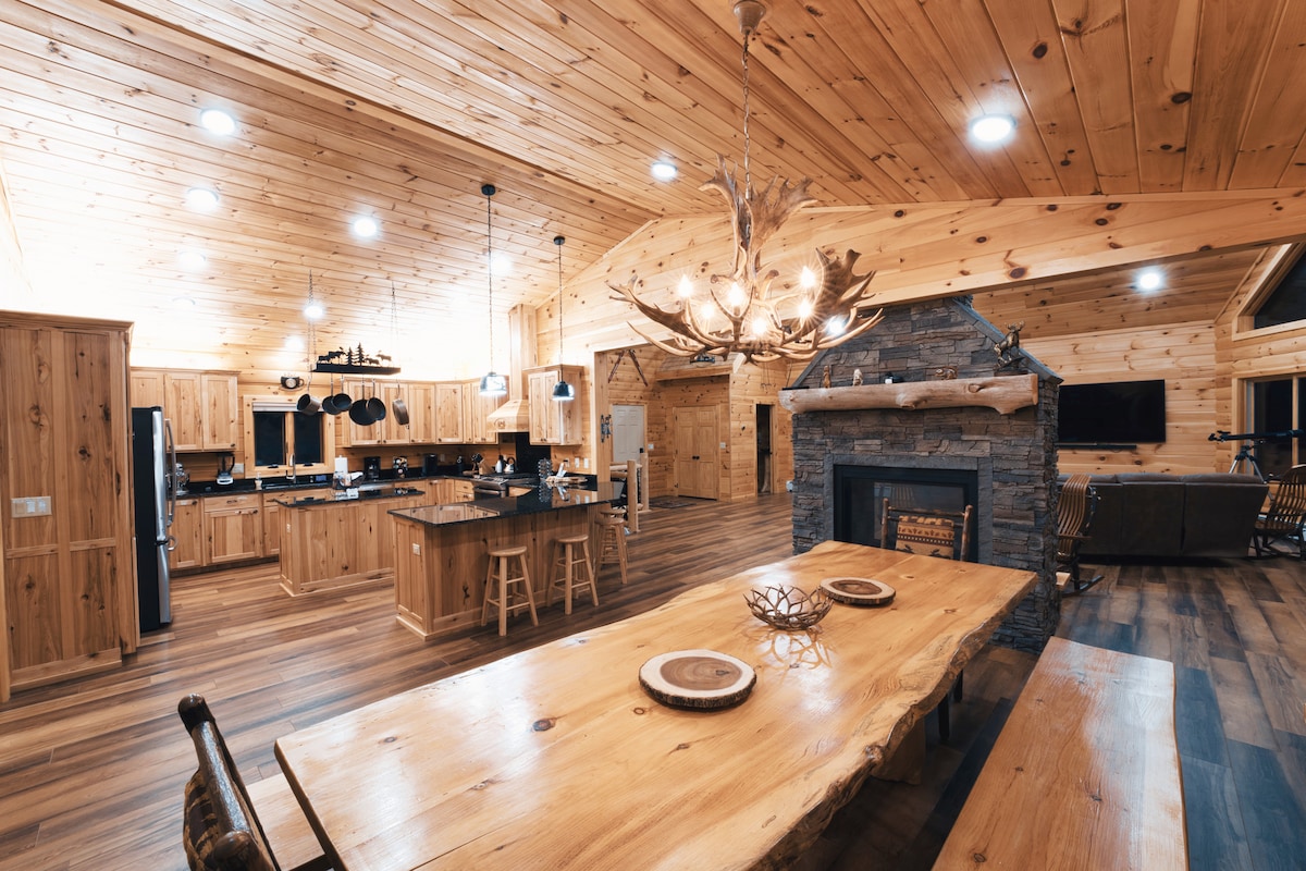 An open-concept kitchen and dining area features warm wooden cabinetry and a large dining table crafted from natural wood. A stone fireplace is centered in the room beneath rustic lighting, enhancing the cozy ambiance. The space is accented with modern appliances and comfortable seating.