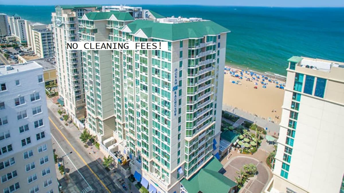 Aerial view of a beachfront resort, showcasing multiple high-rise buildings with green roofs. The vibrant blue ocean is visible alongside a sandy beach populated with colorful umbrellas and beachgoers. The bustling beachfront street is lined with palm trees and amenities.