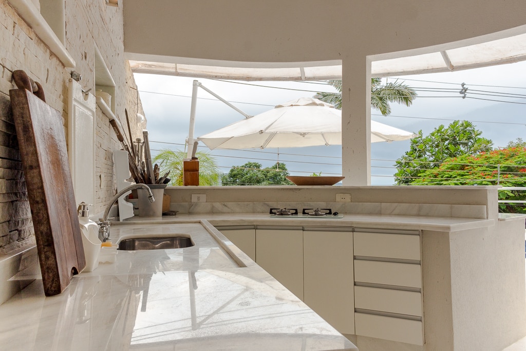 The open kitchen features a curved marble countertop and modern cabinetry, providing ample workspace. Natural light enters through large windows, revealing views of the outdoors and a patio umbrella. A gas cooktop is positioned for convenience, enhancing the inviting design.