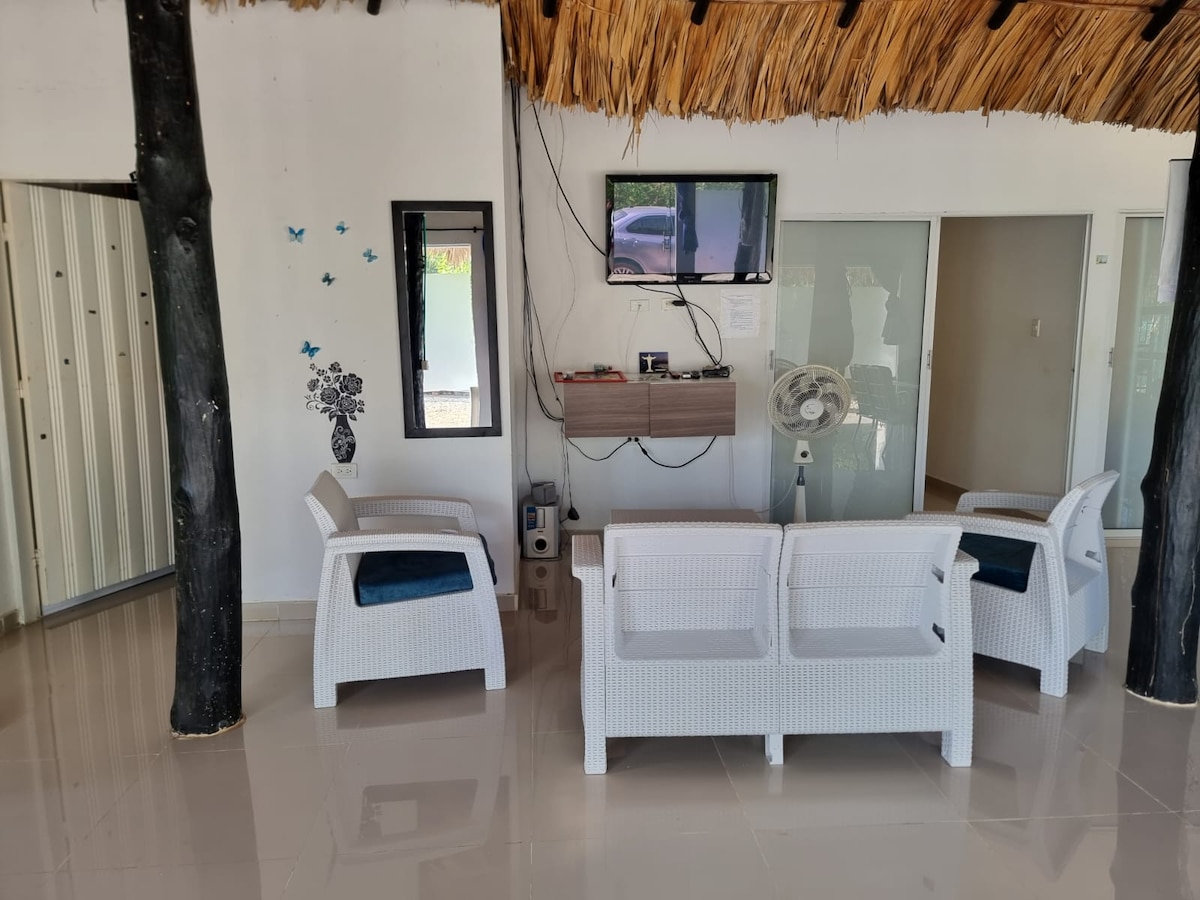 A spacious seating area features four white wicker chairs arranged around a minimalist wooden table. A flat-screen television is mounted on the wall, and a fan is positioned nearby. Natural light enters through a large window, enhancing the airy atmosphere.
