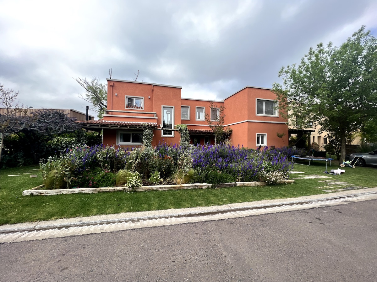 The exterior of the house is presented, featuring a two-story structure adorned with vibrant orange walls. Lush gardens filled with purple flowers and greenery surround the home, while a pathway of stone borders the well-maintained lawn. A tranquil atmosphere is suggested by the cloudy sky.
