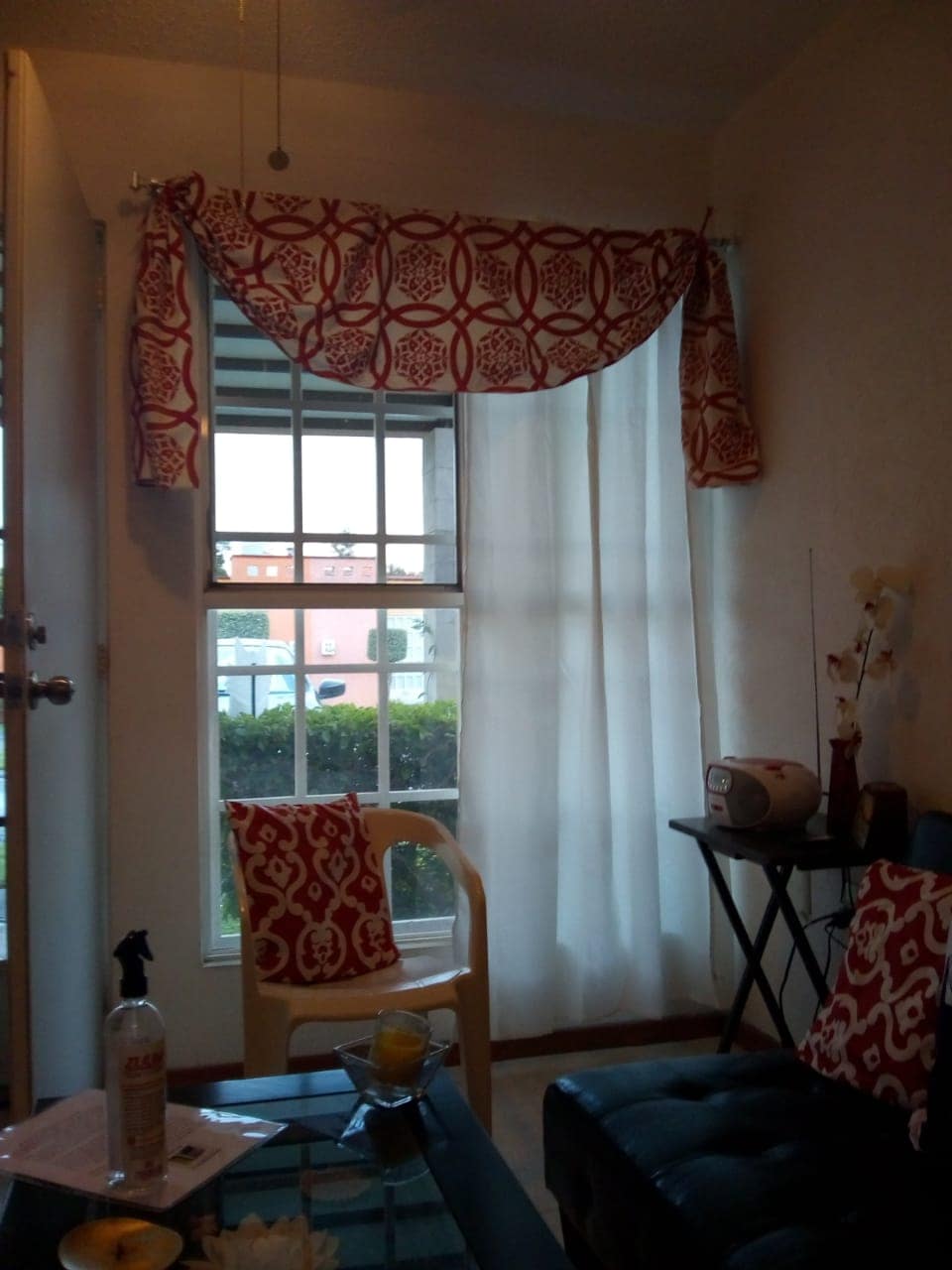 A window with sheer white curtains allows natural light to brighten the room, complemented by decorative red curtains. A small black table and a yellow chair are present, along with patterned cushions, creating a comfortable and welcoming seating area. Outdoor greenery is visible through the window.