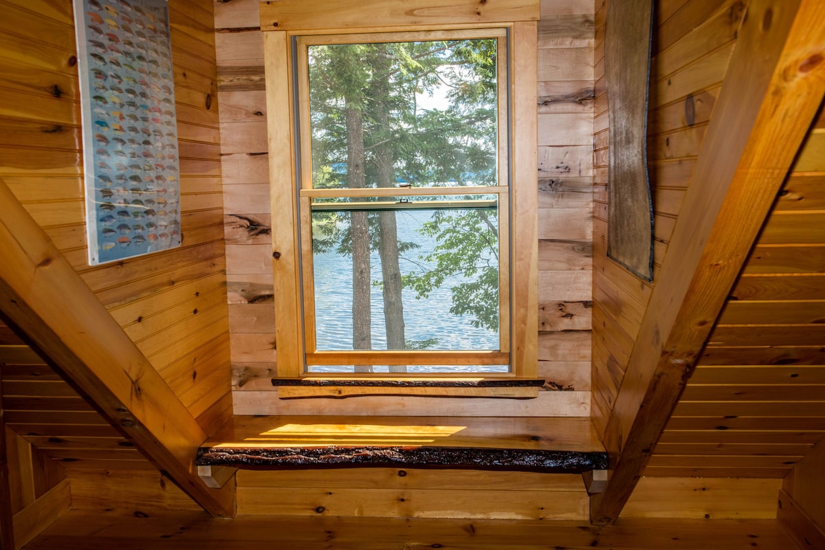 A window framed by wooden paneling provides a view of Webb Lake and surrounding trees. Natural sunlight illuminates the indoor space, highlighting the cabin's warm, handcrafted log construction. A rustic wooden ledge rests beneath the window, enhancing the cozy interior ambiance.