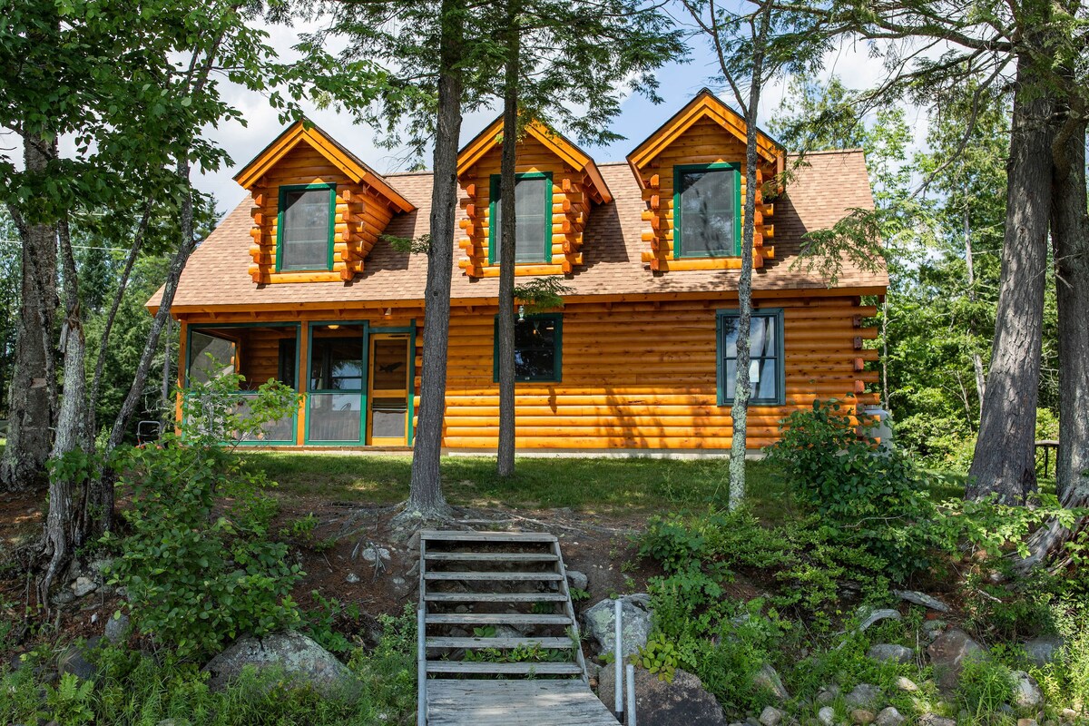 The log cabin exterior features a warm wood finish, complemented by large windows on the upper levels. Flanked by trees, a wooden walkway leads to the entrance, enhancing the connection to nature. The surrounding greenery provides a secluded atmosphere.
