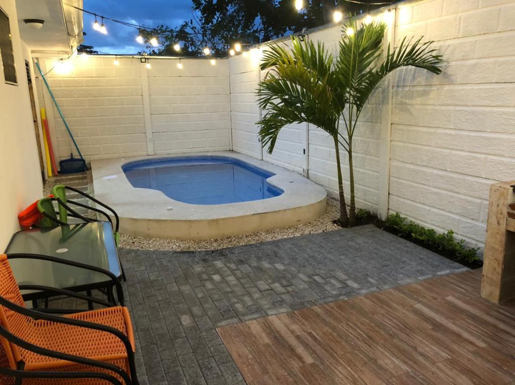 A private pool area is shown, featuring a small blue swimming pool surrounded by well-maintained greenery. Comfortable chairs are positioned nearby on a paved surface, with softly lit string lights hanging above. A clean, white wall provides a serene backdrop.