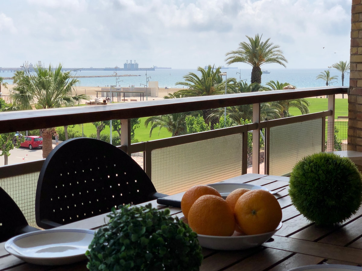 A spacious terrace features a wooden dining table set with plates, vibrant oranges, and green decorative plants. The view includes a serene beach and palm trees, with the blue sea and distant structures visible under a cloudy sky.