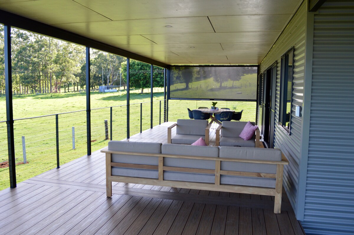 An outdoor deck features a wooden bench with cushions and a small table with chairs. The area offers views of the surrounding fields and trees, creating an open and airy setting that invites relaxation.