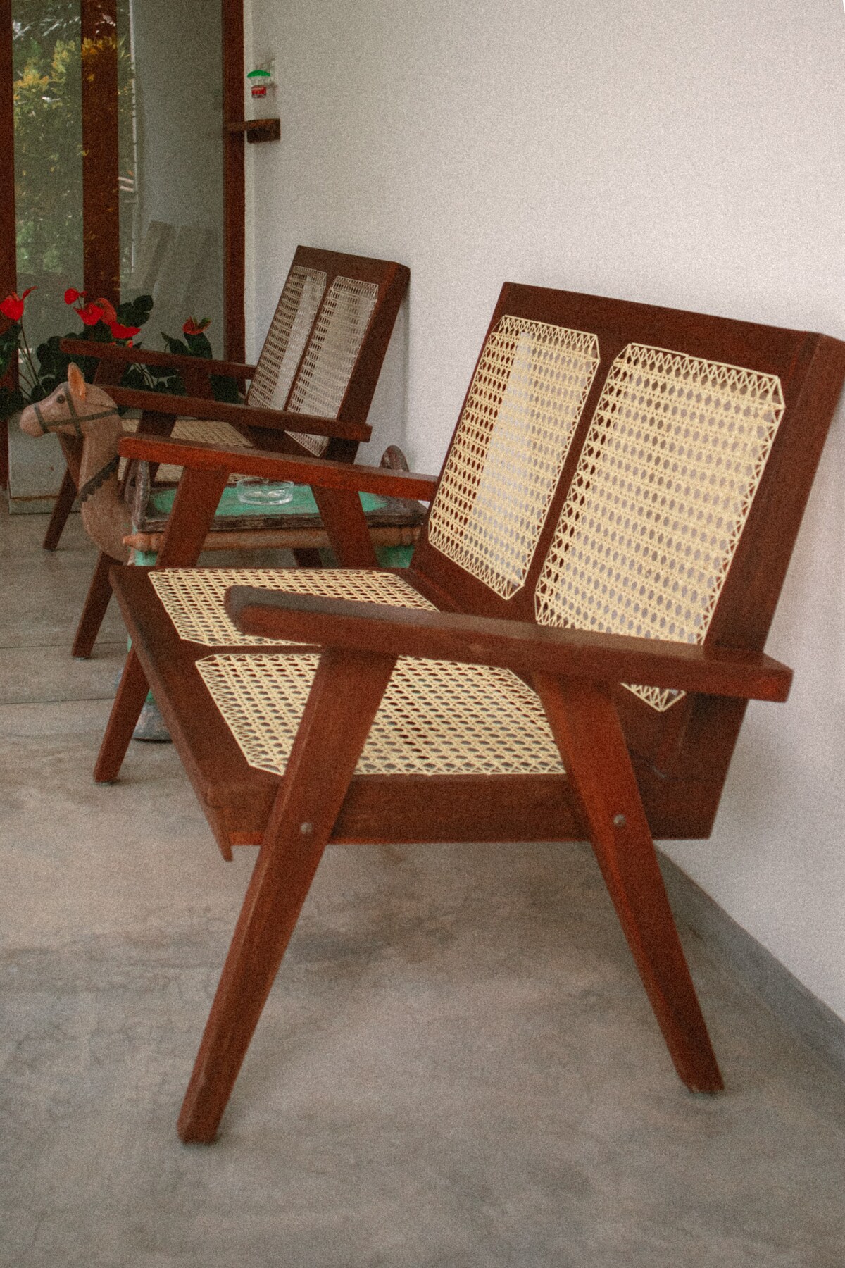 Two wooden chairs are positioned side by side against a light-colored wall. Each chair features woven cane seating and a sturdy wooden frame. A gentle reflection of greenery can be observed in the nearby glass, contributing to a relaxed atmosphere.