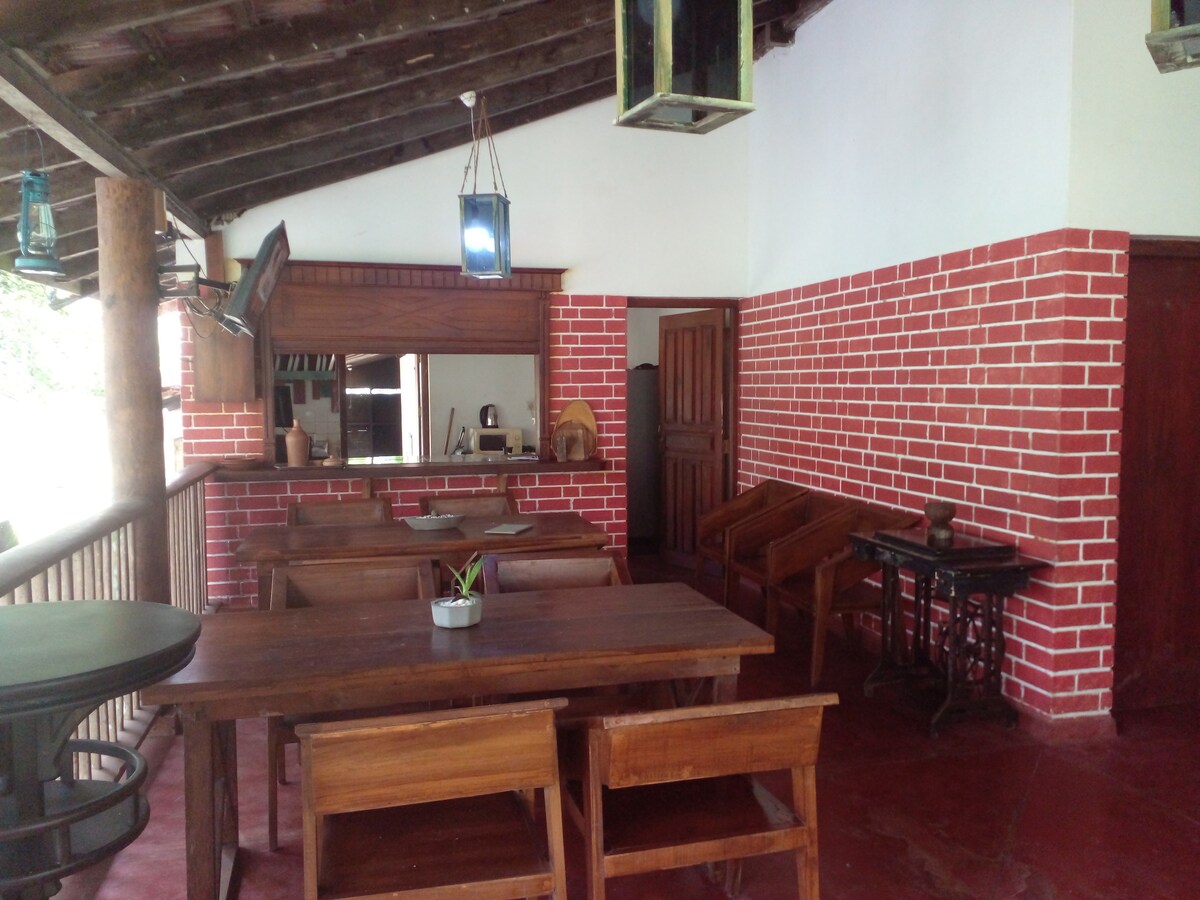An open dining area is framed by red-brick walls and wooden beams. A long wooden table is surrounded by matching chairs. A small potted plant decorates the table's center. Adjacent, a wooden bar counter and partially visible kitchen create a cozy gathering space.