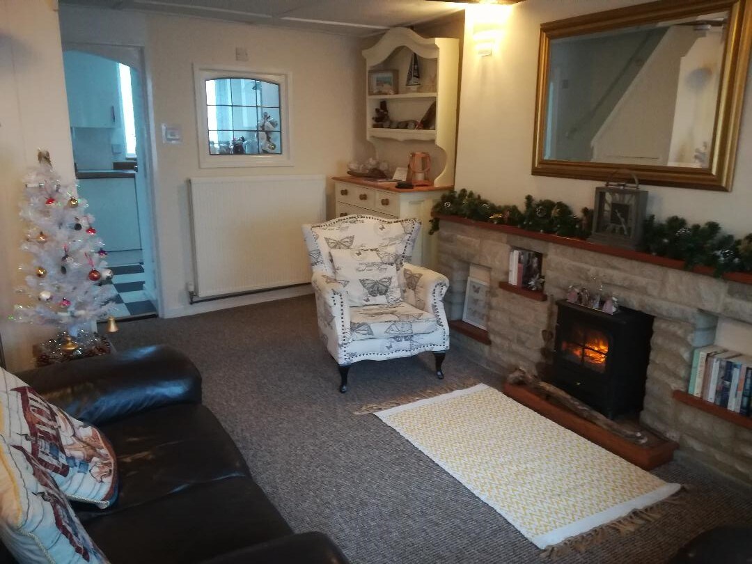 A cozy living area is presented, featuring a plush dark leather sofa and a patterned armchair. A small decorative tree adds a festive touch beside the fireplace, which is set within a stone mantel. Books are neatly arranged on a shelf, contributing to the inviting ambiance.