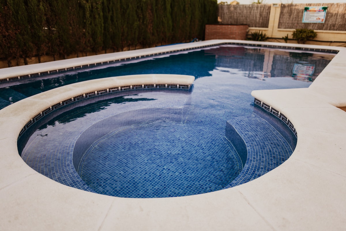 A shared swimming pool is framed by clean white tiles, featuring a rounded area that offers a relaxing space. The deep blue water reflects the surrounding greenery, providing a serene environment for guests. The pool area is enclosed for privacy and comfort.