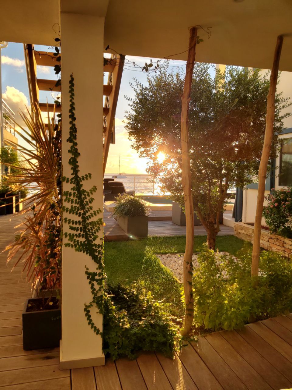 Sunlight filters through foliage onto a manicured garden area. A wooden deck is seen in the background, with the ocean visible beyond. Staircases lead to elevated spaces, surrounded by plants that enhance the serene atmosphere.
