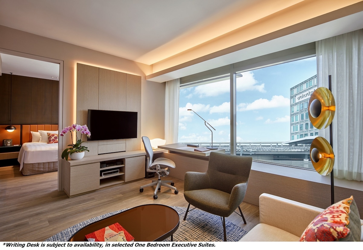 A modern living space showcases large windows allowing natural light and city views. A flat-screen television is mounted on a wood-paneled wall above a low-profile entertainment unit. A comfortable chair and writing desk are positioned near the window, creating a functional area.