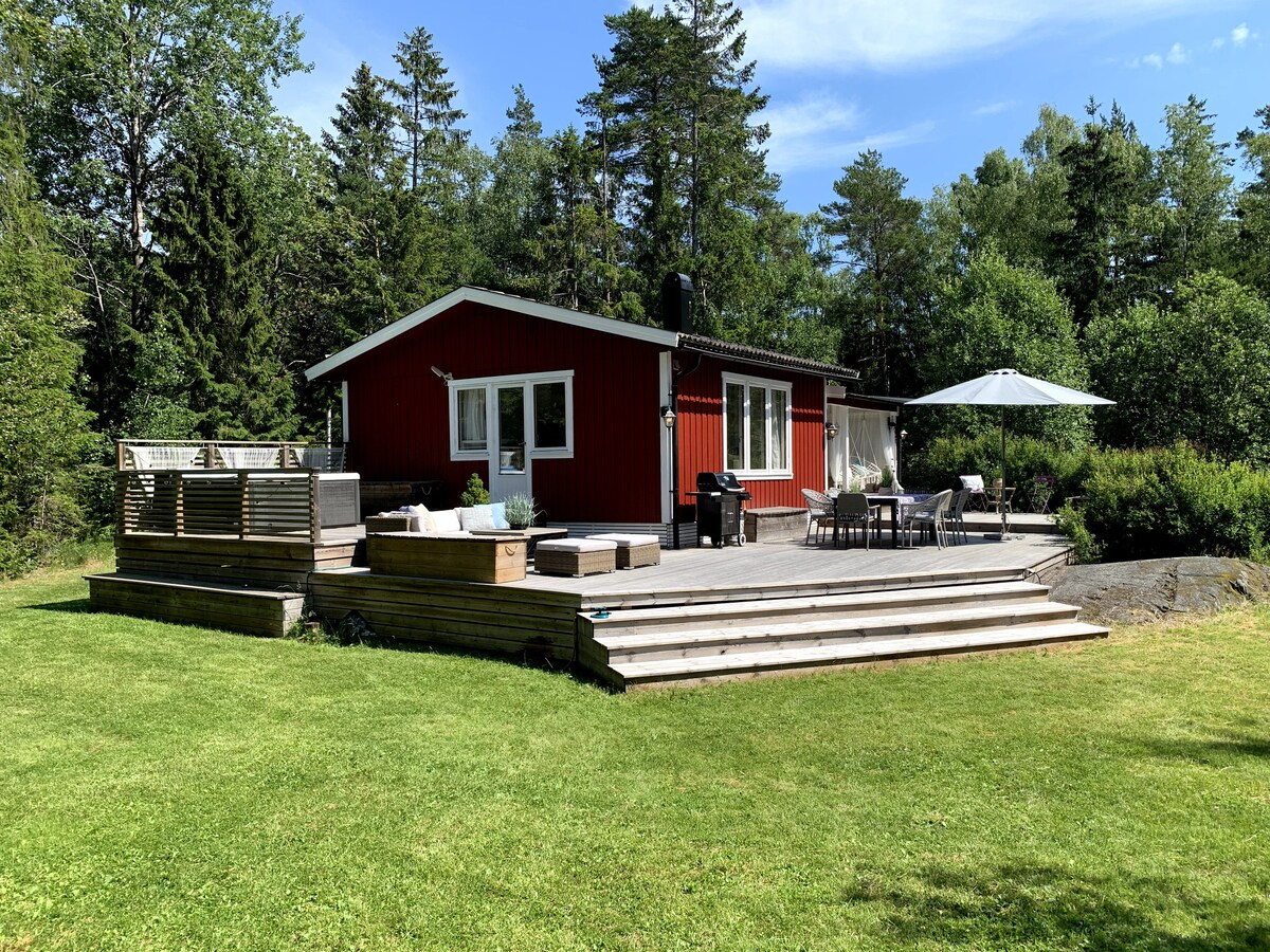A red cottage is positioned amidst greenery, featuring a spacious wooden deck with seating and a large umbrella. The outdoor space includes a grill and steps leading to a grassy area, enhancing the connection with nature.