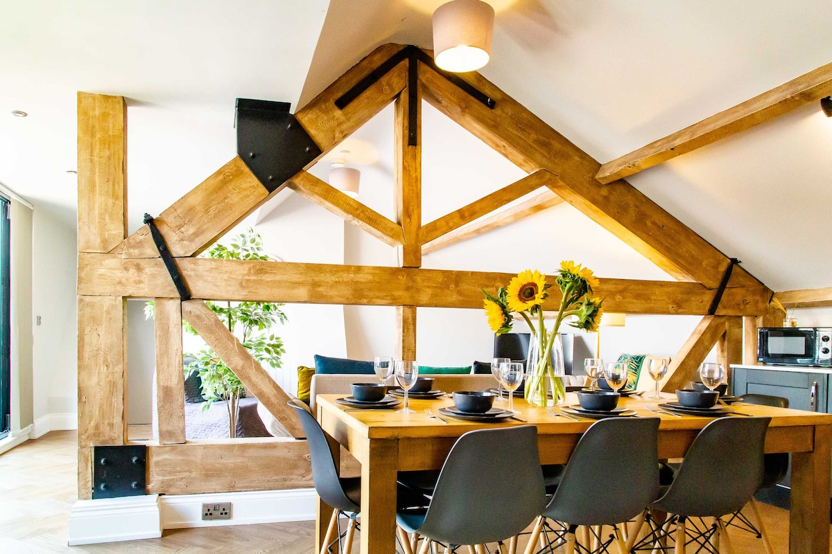 A tastefully arranged dining area is highlighted by a large wooden table, surrounded by sleek black chairs. Sunflowers in a vase add a touch of warmth. Original wooden beams and large glass doors enhance the contemporary design, connecting the space to the outdoors.
