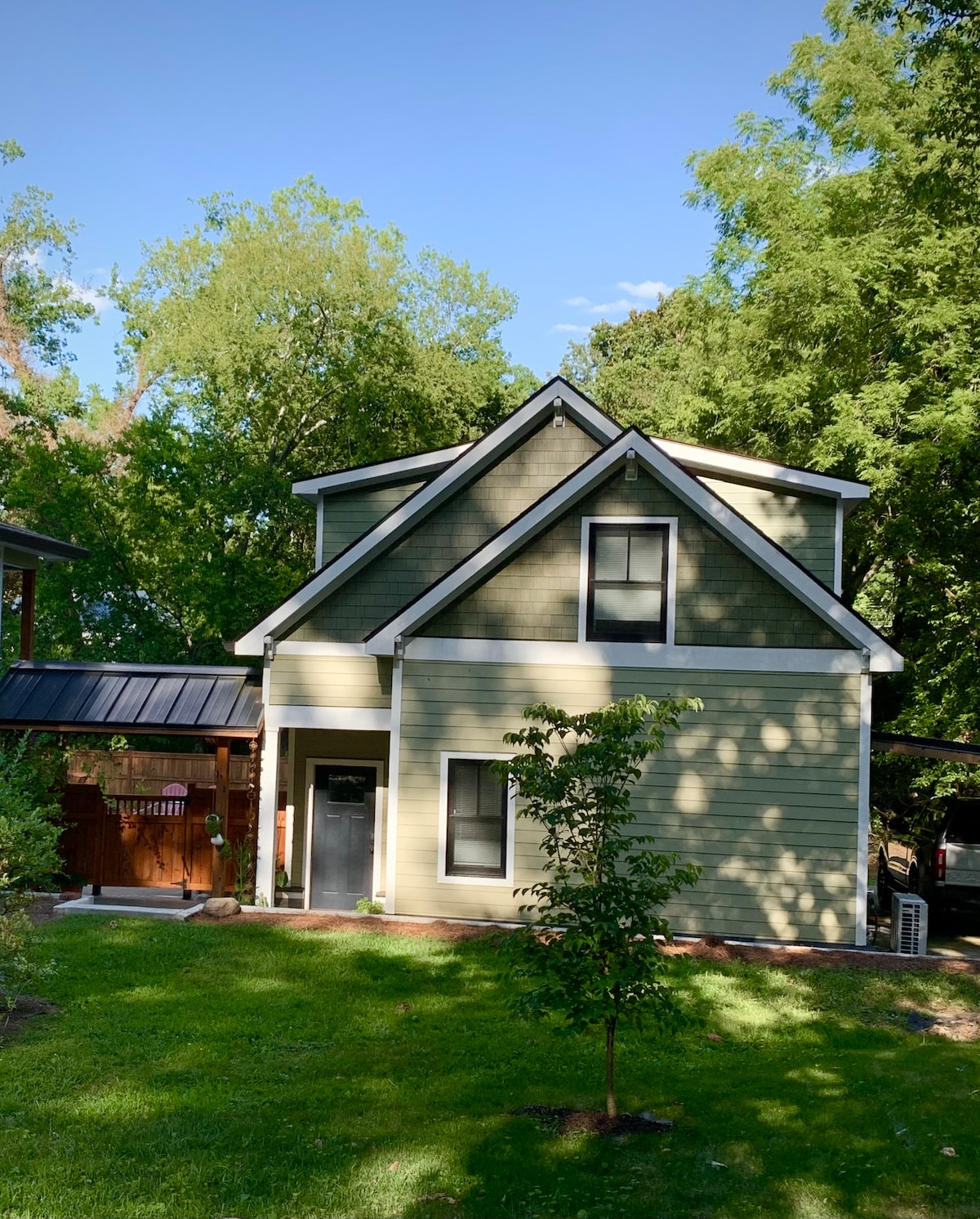 The exterior of a two-story house is depicted, showcasing green siding and a black metal roof. A well-maintained lawn surrounds the home, with several trees providing shade. A separate entrance is visible, emphasizing accessibility. The neighboring structures add to the residential ambiance.