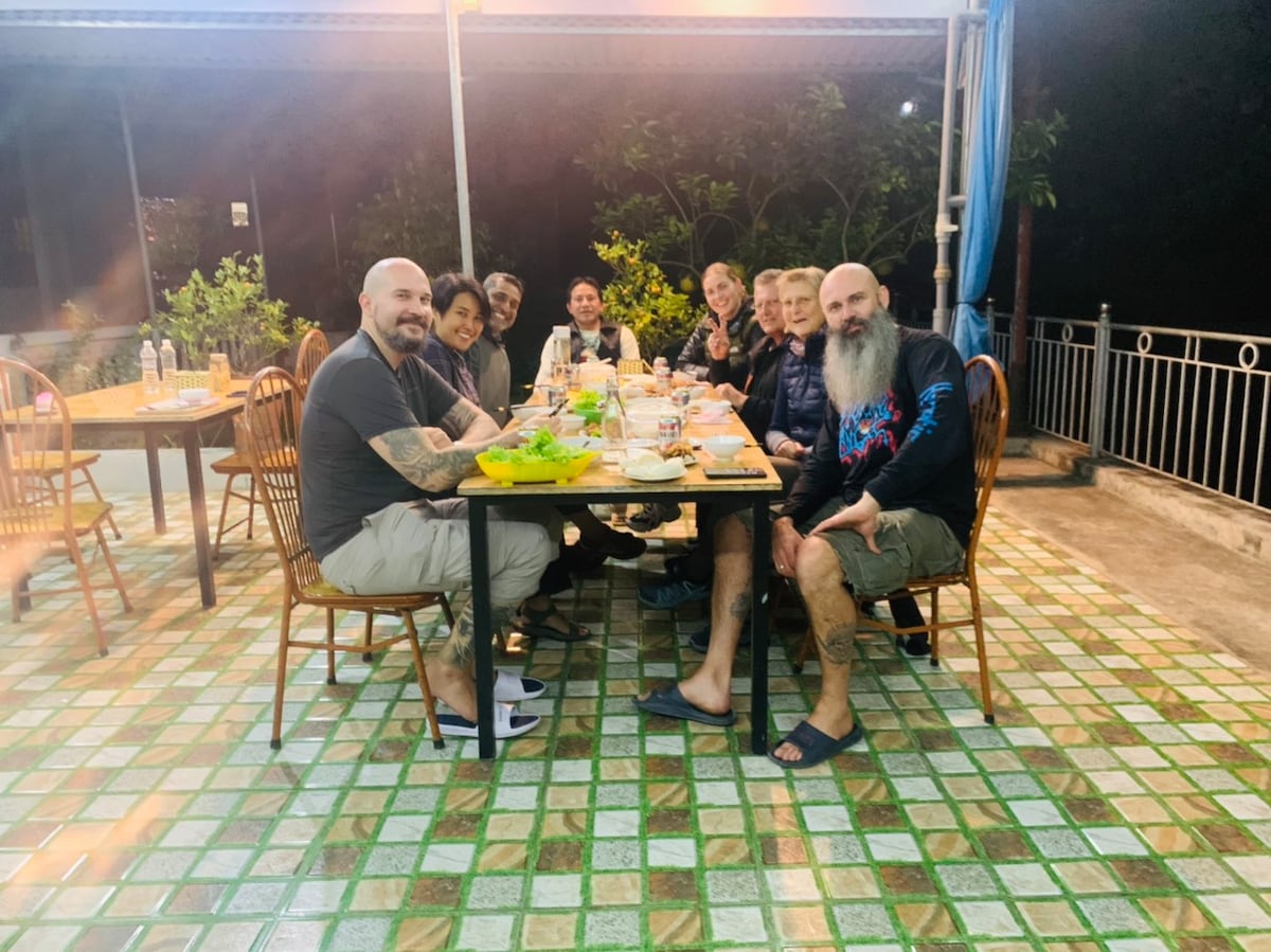 A dining area is shown with a long table set for a meal, surrounded by several chairs. The setting is illuminated by soft lighting, with greenery visible in the background, contributing to a relaxed ambiance. The floor features a patterned tile design.