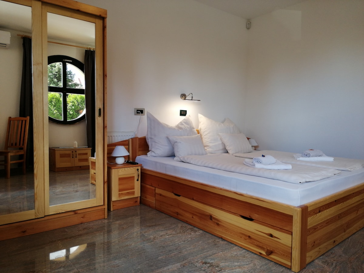 The sleeping area features a wooden bed with white linens and multiple pillows. A circular window allows natural light to enter. Nightstands with lamps are positioned on either side of the bed, and a mirrored wardrobe is visible in the background.
