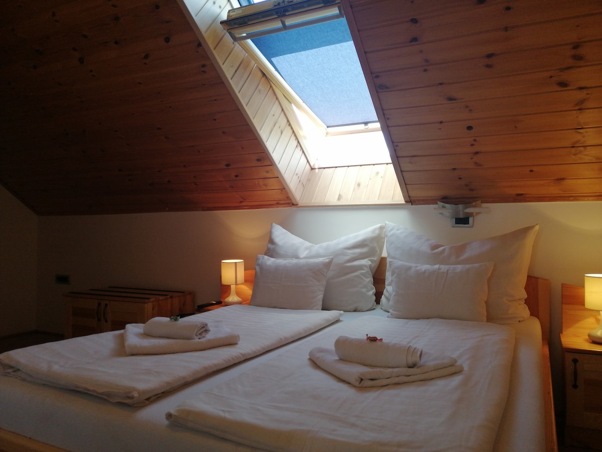 The bedroom features two spacious beds with white linens and neatly arranged towels. Soft lighting from bedside lamps enhances the warm wood paneling of the ceiling, while a skylight allows natural light to fill the space.