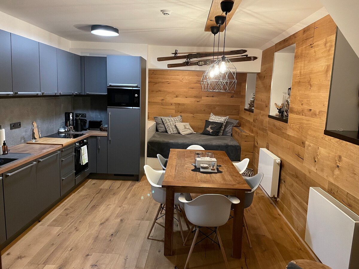 The image displays a modern kitchen area featuring sleek gray cabinets and a compact dining table surrounded by white chairs. A cozy seating nook with decorative pillows is situated adjacent to the kitchen, all enhanced by warm wooden paneling and contemporary lighting fixtures.