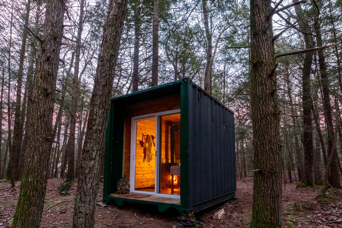 A shipping container cabin is situated among tall trees, with warm light glowing from its interior. Large windows frame a view of the forest, inviting the natural surroundings into the space. The rustic exterior blends with the woodland setting, providing a sense of seclusion.