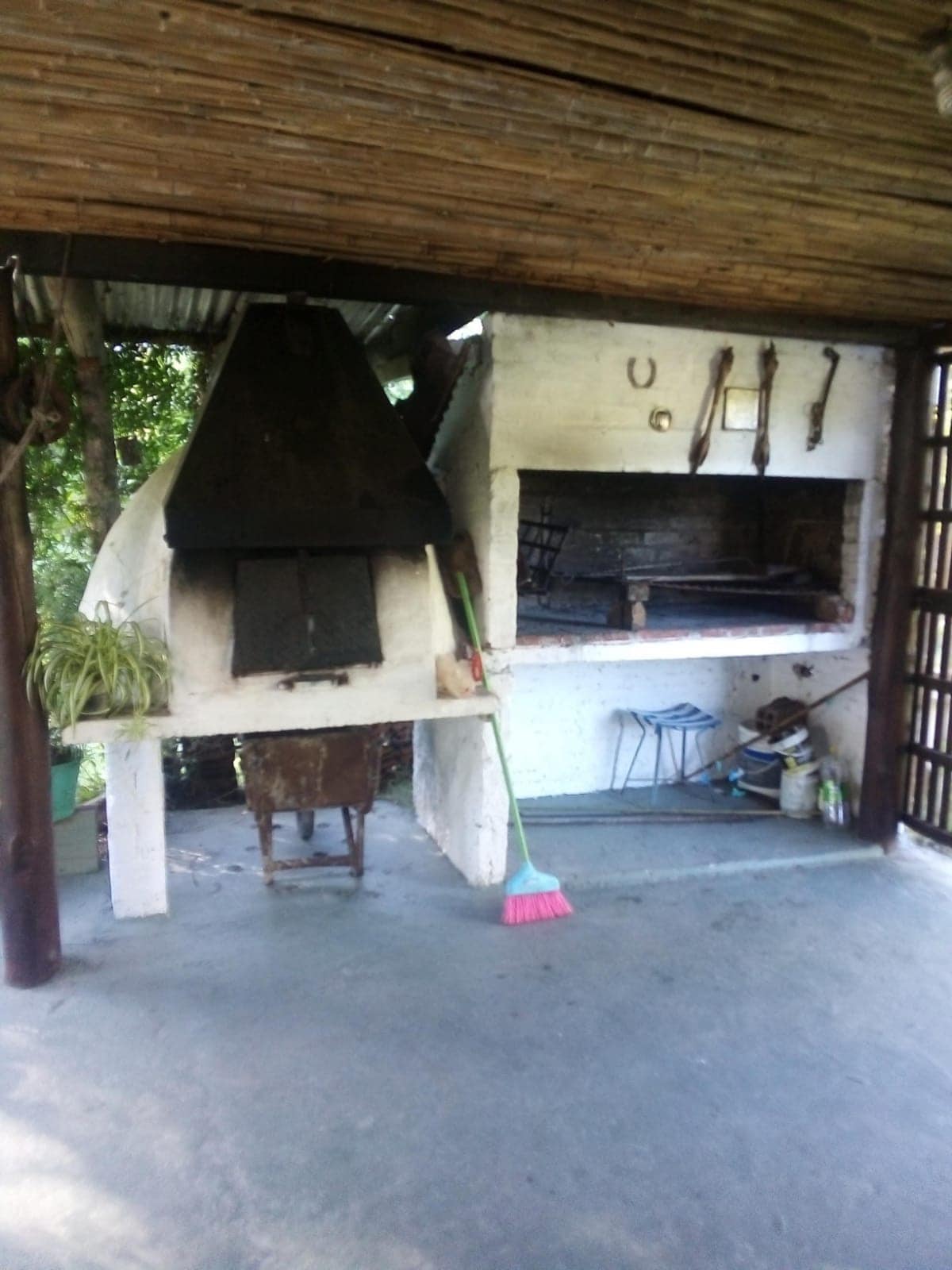 An outdoor grilling area is featured, with a built-in barbecue on one side and a traditional wood-fired oven on the other. The space is equipped with a broom leaning against the wall and is surrounded by greenery, creating a relaxing environment ideal for cooking and gathering.