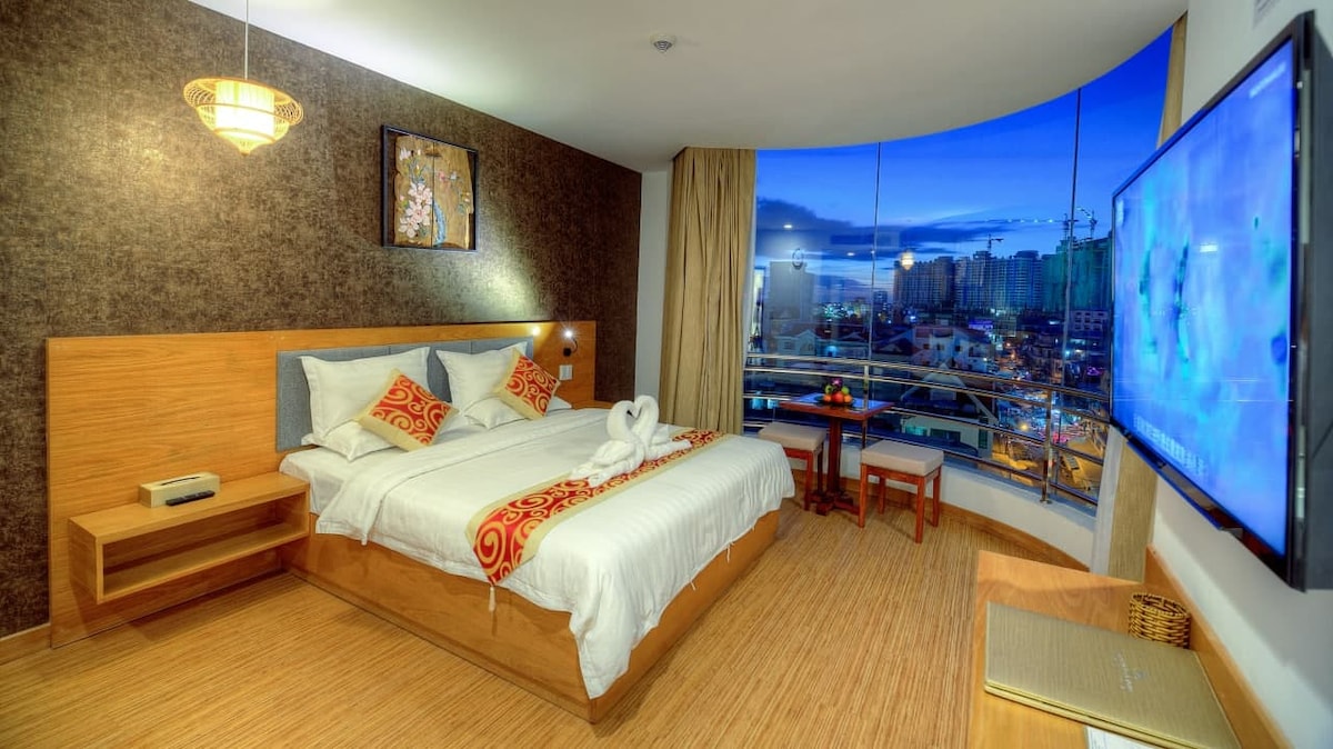 A spacious bedroom features a large bed with decorative pillows and a plush white blanket. Natural light streams through expansive windows, offering views of the city skyline. A flat-screen television is mounted on the wall, and wooden accents provide a warm touch to the modern design.
