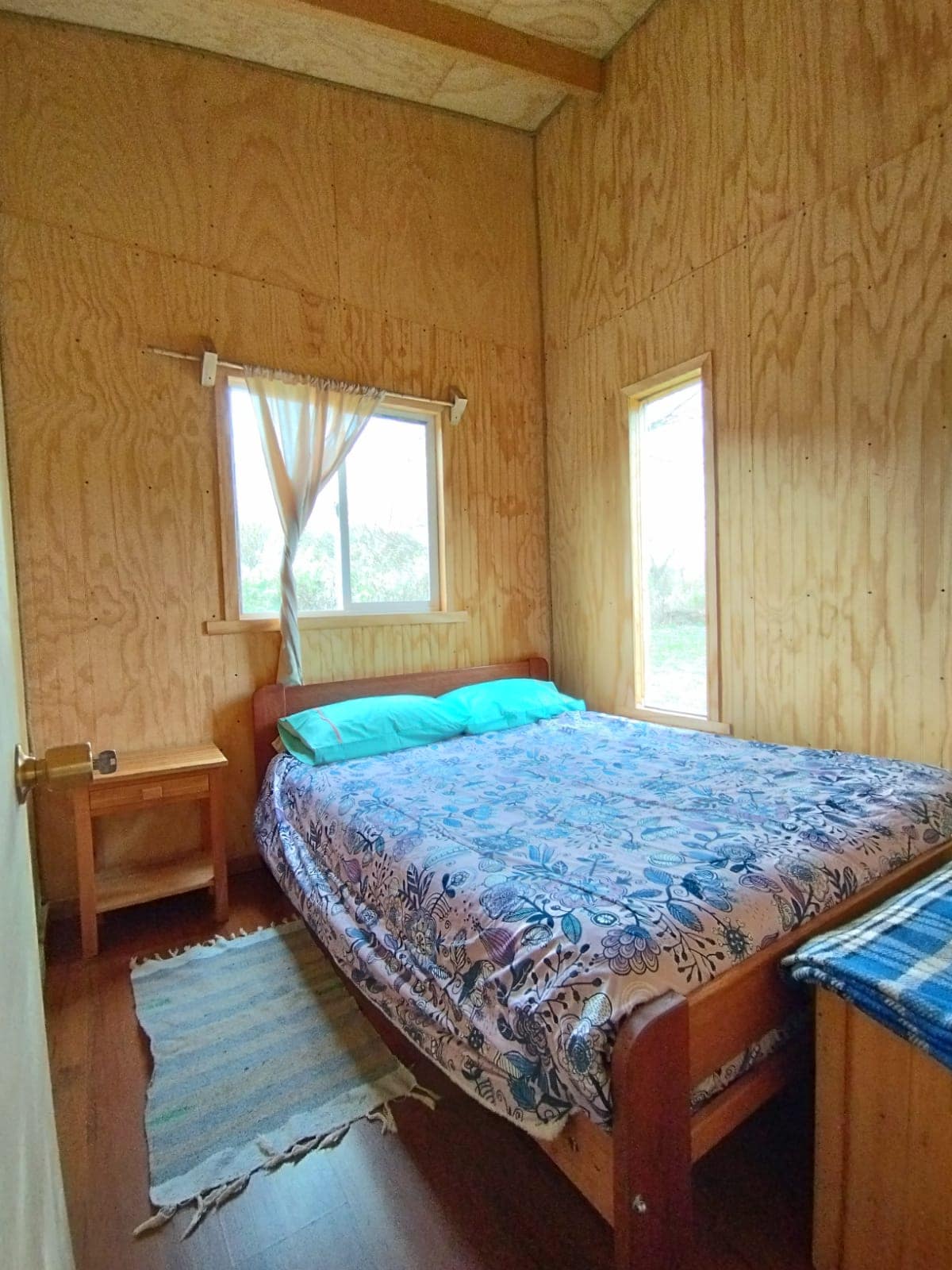 A cozy sleeping area features a spacious bed dressed with a floral-patterned blanket and blue pillows. Two windows allow natural light to fill the room, complemented by wooden walls and flooring. A small nightstand is positioned beside the bed, enhancing the simplicity of the space.