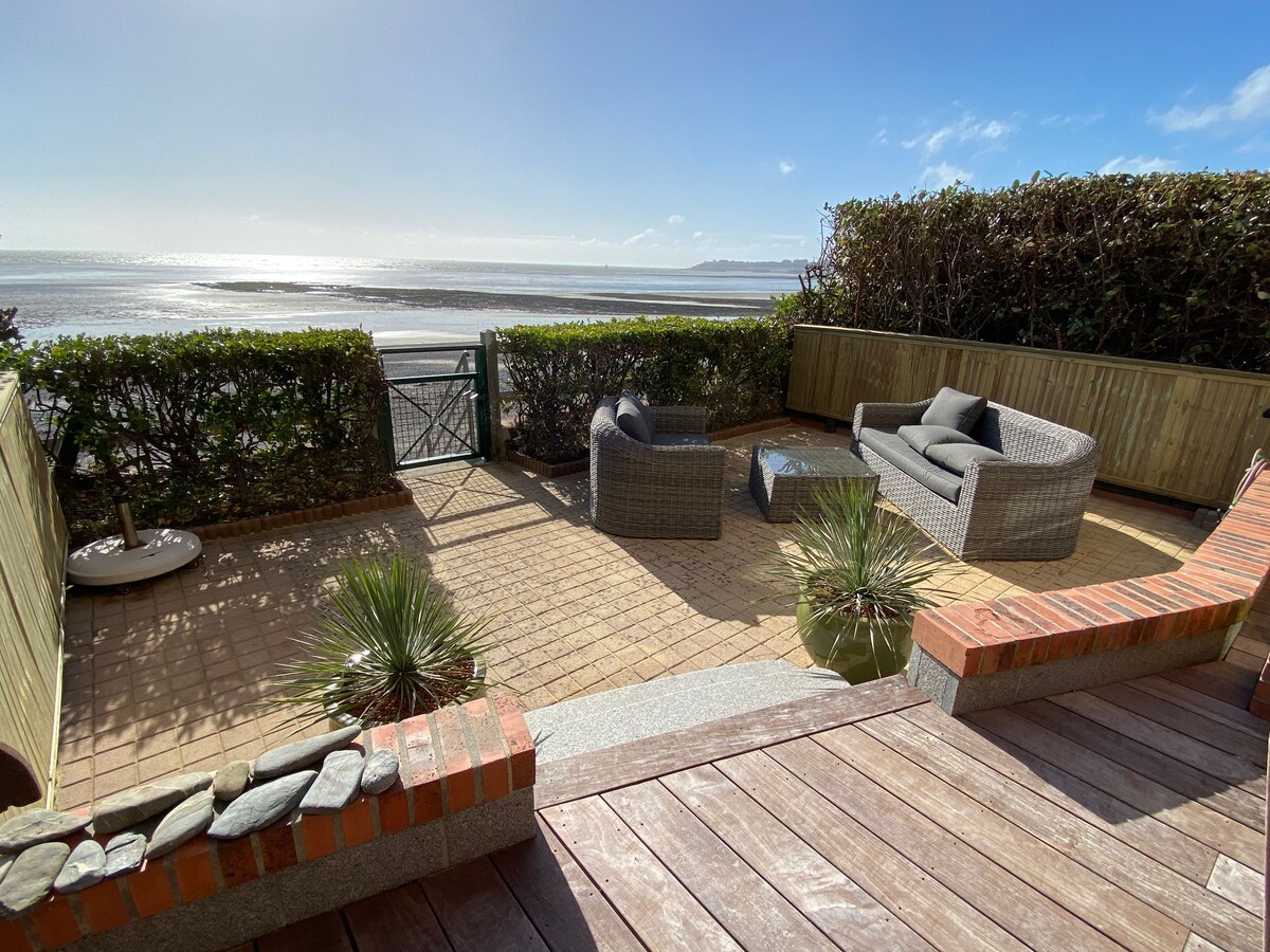 A terrace area is presented with outdoor seating arrangements, including comfortable woven furniture. Plants and low hedges create a boundary, while a pathway leads to a gate that opens towards the beach. Views of the sea extend beyond the terrace, under a clear sky.