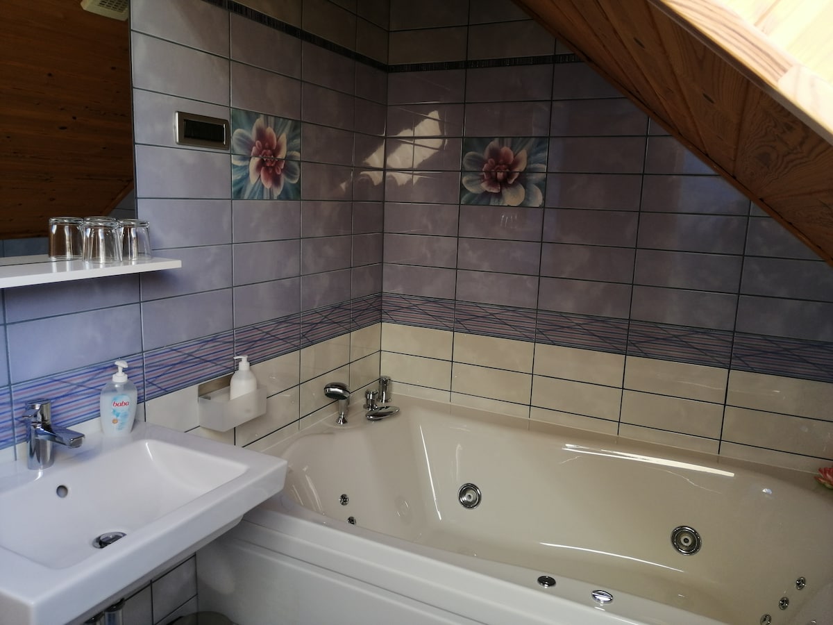 A spacious bathroom features a whirlpool tub against decorative tile walls adorned with floral patterns. A modern sink is positioned near the tub, with toiletries and drinking glasses neatly arranged on a shelf. Natural light highlights the unique ceiling design.