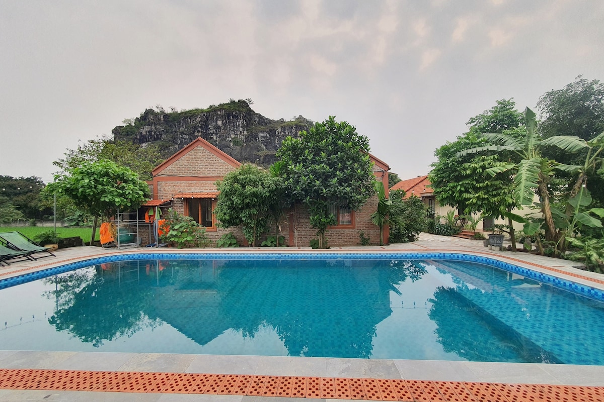 A serene outdoor pool surrounded by lush greenery is visible, with a backdrop of a rocky hill. The pool’s clear blue water reflects the soft hues of the sky, while comfortable lounge chairs are placed nearby for relaxation.