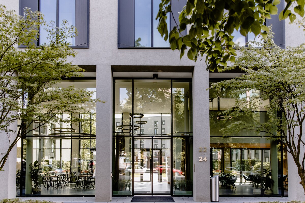 The modern building entrance features large glass doors framed by sleek gray walls. Symmetrical outdoor seating is arranged in a shaded area, complemented by strategically placed trees that add greenery to the surroundings.