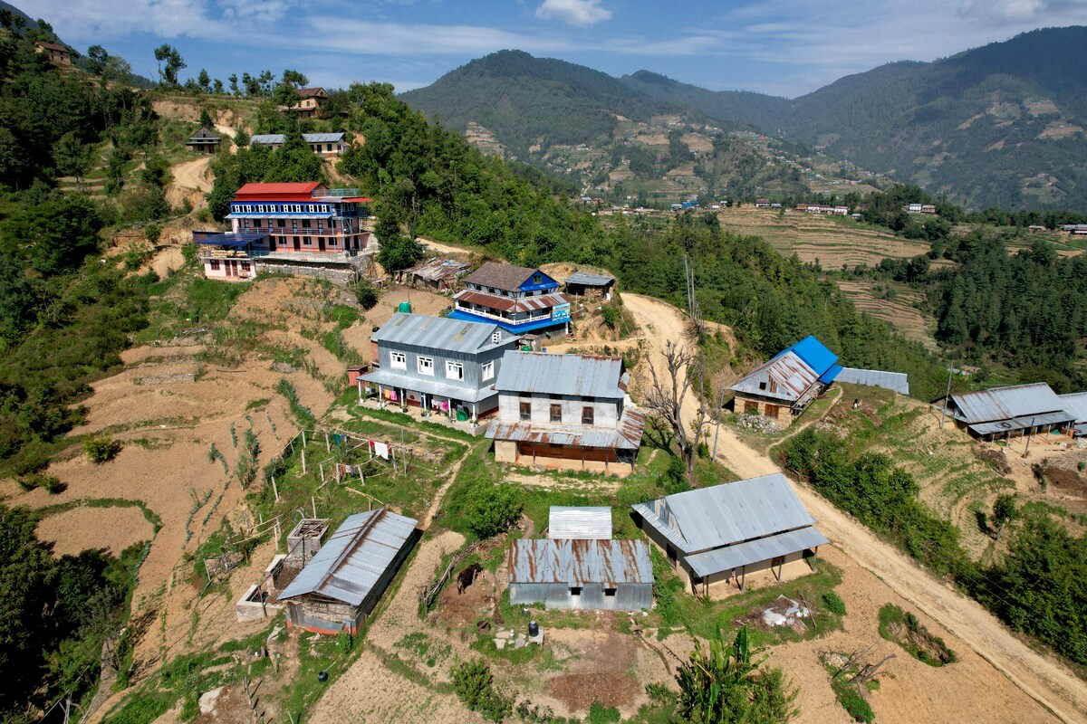 An aerial view captures a charming village nestled in the hills, framed by greenery and agricultural land. Various buildings with metal roofs are visible, interspersed among cultivated fields and trees. The surrounding mountains create a serene backdrop under a clear blue sky.
