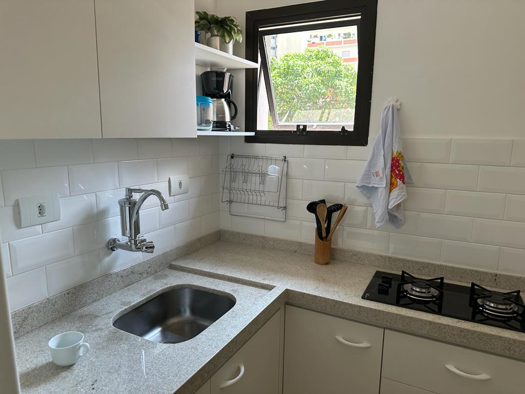 A compact kitchen features modern cabinetry and a granite countertop. A stainless steel sink and gas stove are situated against a white tiled backsplash. Natural light enters through a window, illuminating a coffee maker, utensils, and a dish rack neatly arranged within the space.