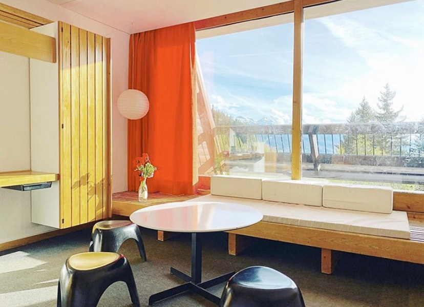 A sunlit living area features large windows showcasing mountain views. A circular white table is accompanied by two black stools. A low bench offers additional seating beside a vibrant orange curtain, while a vase of flowers adds a touch of color to the space.