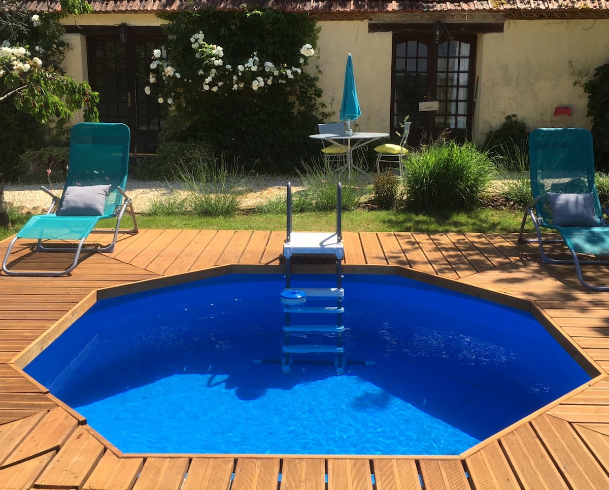 An octagonal private pool is surrounded by a wooden deck, featuring two lounge chairs and a small table set in the background. Lush greenery adds a touch of nature, while doors visible on the cottage hint at inviting indoor spaces.