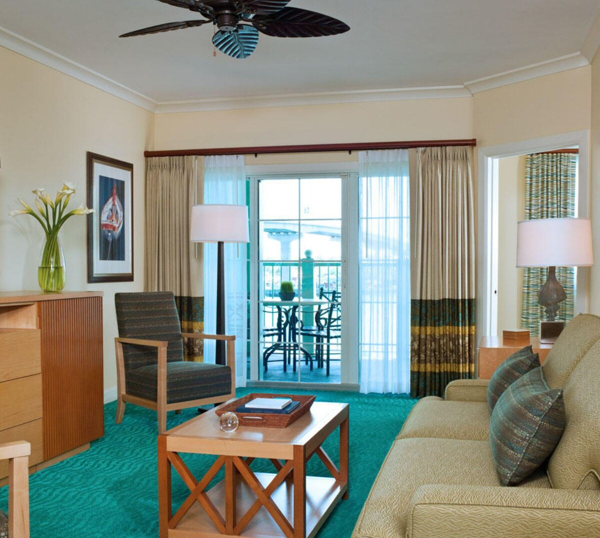 A welcoming living space features a comfortable sofa and an armchair beside a wooden coffee table. Large sliding glass doors open to a balcony, allowing natural light to enhance the teal carpet. Soft curtains frame the windows, and decorative elements include a plant and framed artwork.