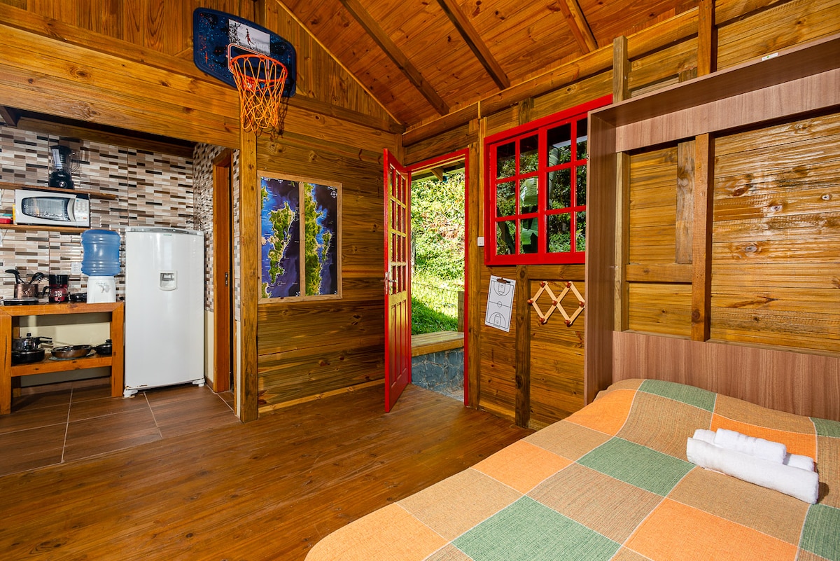 A wooden interior showcases a cozy sleeping area with a colorful blanket draped over the bed. Bright red doors open to a green landscape outside, while a small kitchenette with a refrigerator and cooking appliances is visible. Basketball hoop decorates the space.