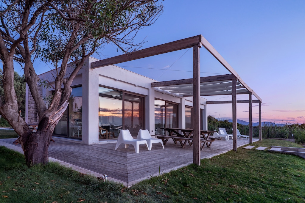 A modern seaside house is shown in twilight, featuring large glass walls that offer expansive views. A wooden patio is furnished with a picnic table and white lounge chairs, surrounded by a lush garden. The structure is partially shaded by a wooden overhang.