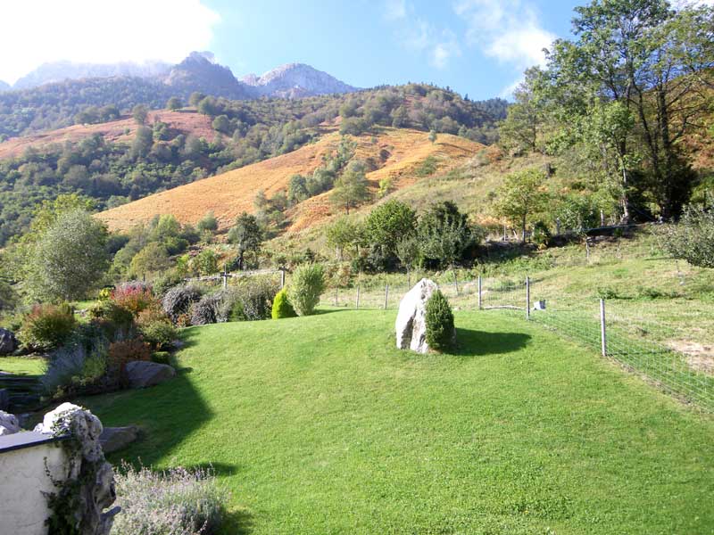 A green landscape is presented, featuring rolling hills and a rocky outcropping. The lawn is well-maintained, surrounded by various trees and shrubs. The backdrop showcases a mountainous terrain with a gradient of autumn colors on the slopes, under a partly cloudy sky.