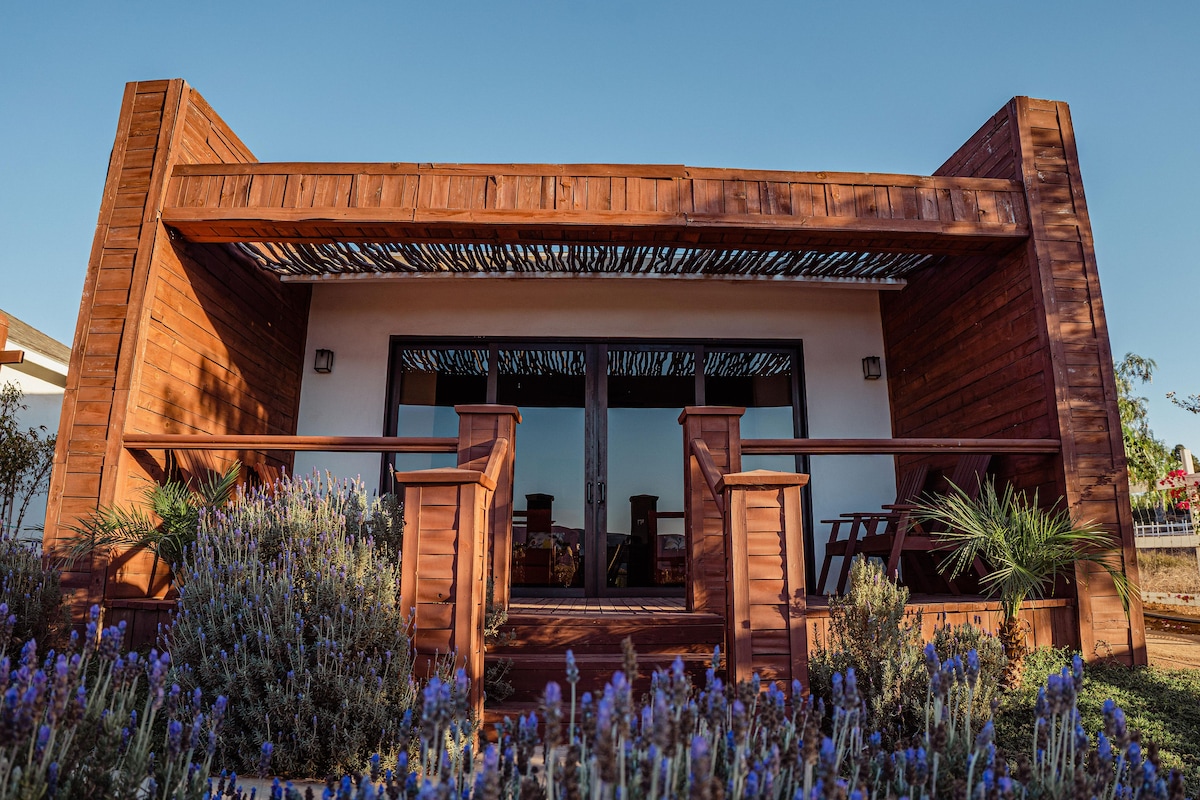 A wooden exterior features a cozy terrace, accented by a row of lavender plants. Large glass doors provide a glimpse into the inviting interior. The structure is framed by wooden beams, enhancing its natural appeal, set against a clear blue sky.