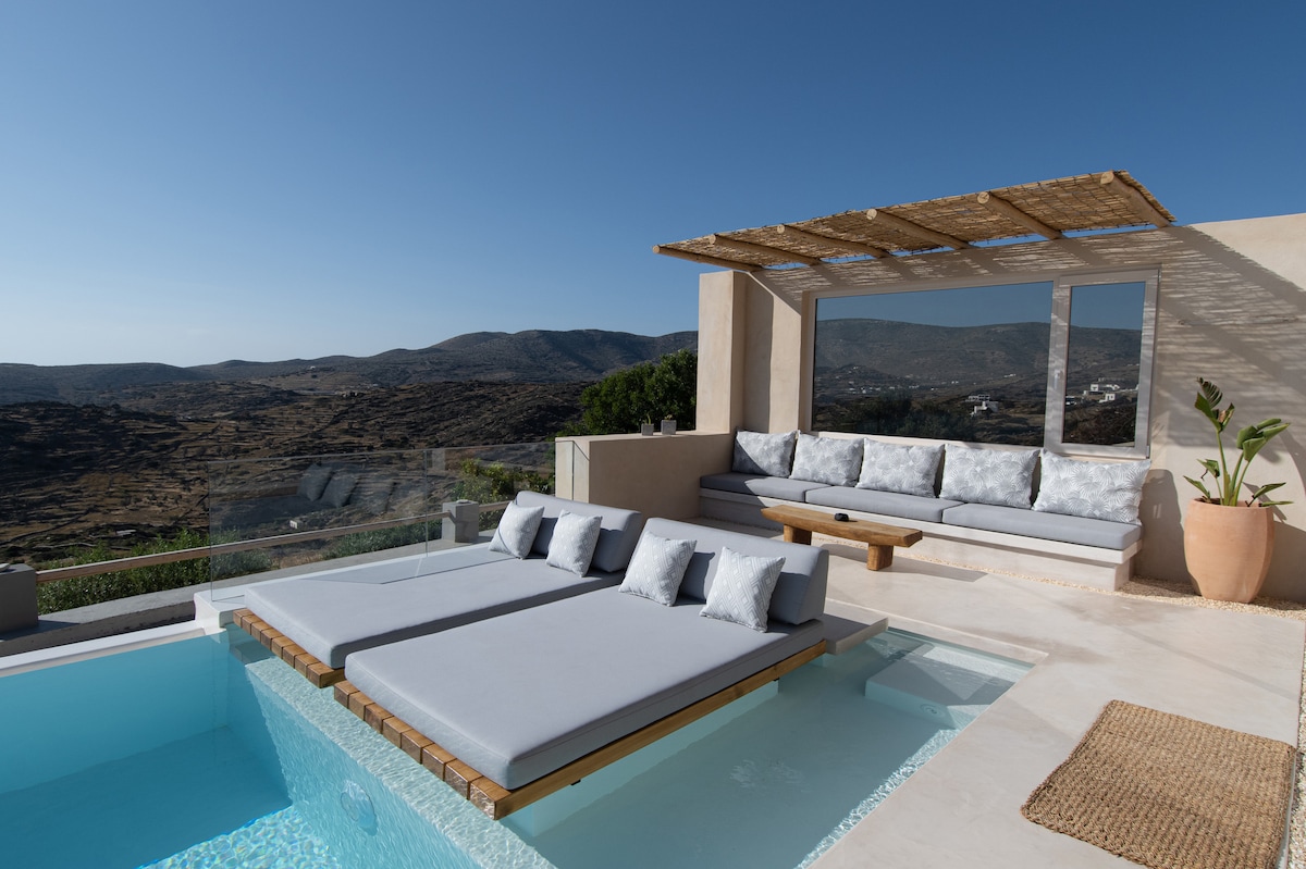 An outdoor area features a private pool with adjacent sun loungers covered in soft grey cushions. A shaded seating nook with light-colored cushions provides views of the surrounding hills, while a potted plant adds a touch of greenery to the space.