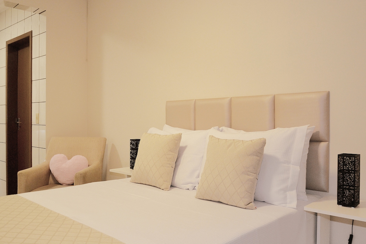 A light-colored room features a spacious bed adorned with soft white bedding and decorative pillows. A cozy armchair sits nearby, complemented by two black bedside lamps. Faint natural light filters in, creating a serene and peaceful ambiance.