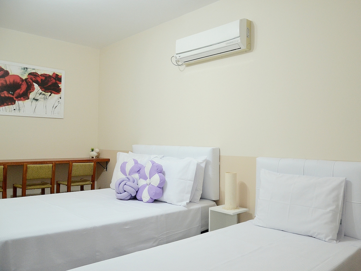The room features two spacious beds with white linens and decorative pillows. A small table with two chairs is visible against the wall, while an air conditioning unit is installed above. Light-colored walls enhance the room's bright and airy feel.
