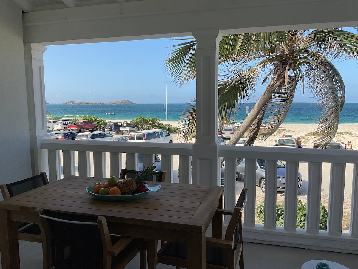 A private balcony offers a view of the beach and ocean, framed by palm trees. A wooden dining table is set with a bowl of fruit, while the sandy shore and vehicles parked nearby are visible in the distance.