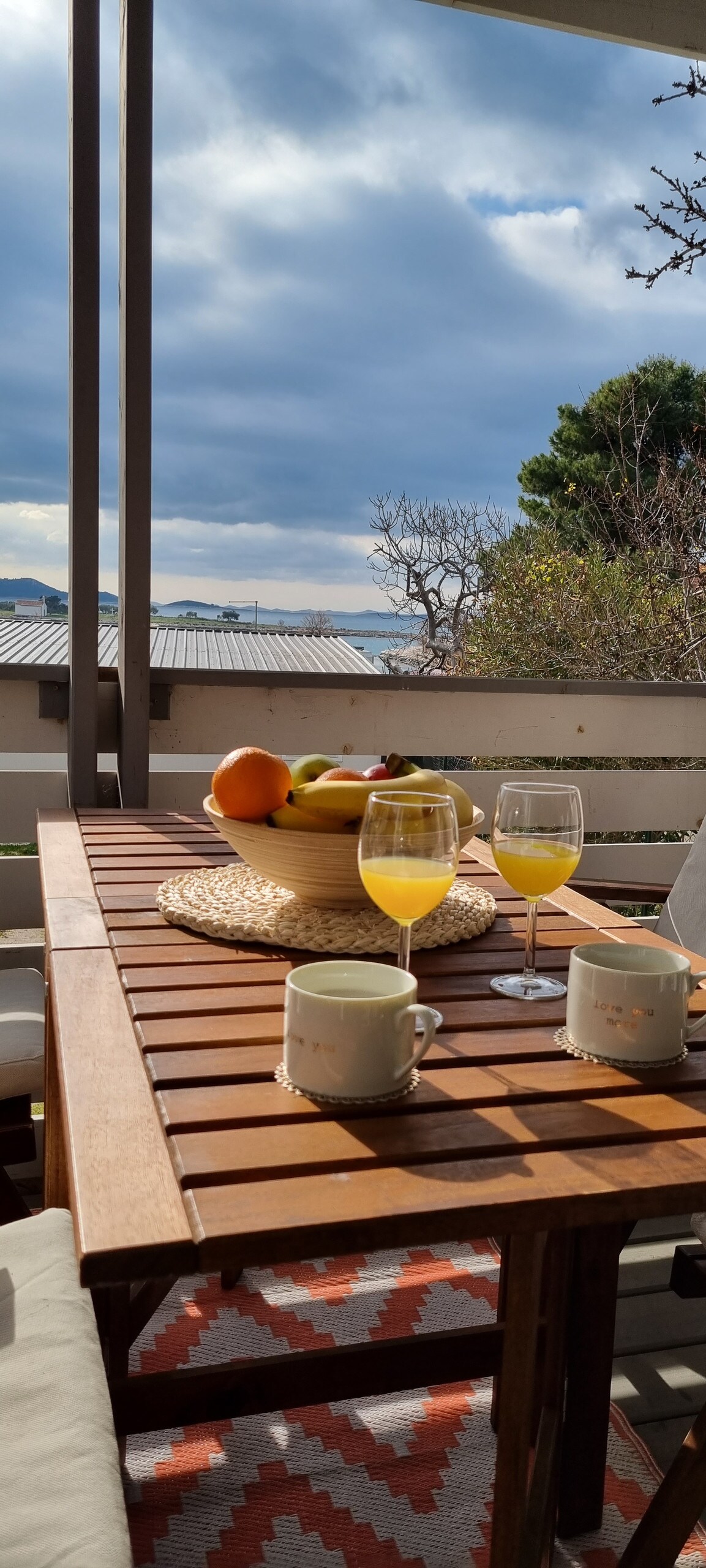 A wooden dining table is set on a terrace, featuring a fruit bowl and two glasses of juice. Coffee mugs rest nearby, with a patterned rug underneath. In the background, a view of the sky and treetops is visible.